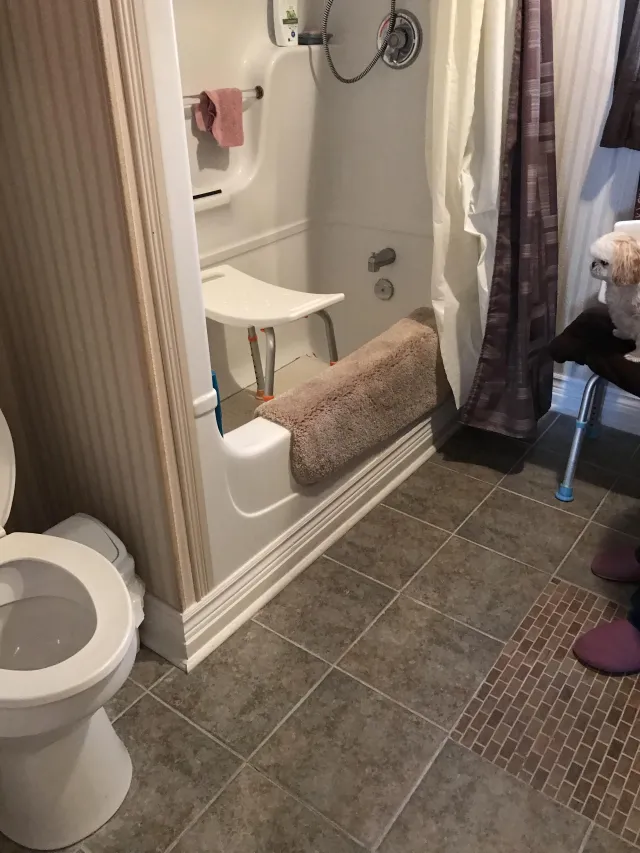 A bathroom with a toilet and a bathtub with a shower seat is shown. The floor is tiled with two different tile patterns. A small white dog is visible in the upper right corner, sitting on a dark surface.