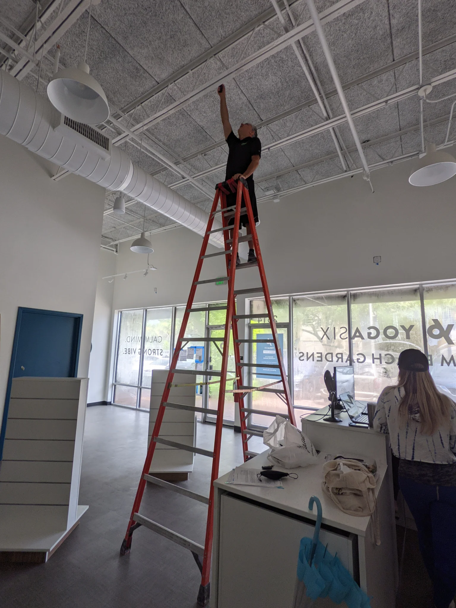A man is standing on a tall red ladder and working on the ceiling lights of a building. There is a woman sitting at a desk in the foreground, and a large blue door on the left side of the image. The building appears to be a yoga studio with "YOGASIX" visible on the window.