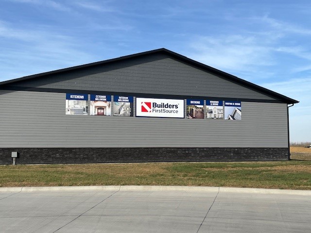 Side view of the Builders FirstSource building in Spencer, showcasing a modern structure with gray horizontal siding and a dark stone accent base. The wall is adorned with multiple promotional panels highlighting various services, such as kitchens, exterior materials, interior millwork, decking, and roofing & siding, all centered around the prominent Builders FirstSource logo. The neatly paved driveway in the foreground contrasts with the green grass lining the building, while a vivid blue sky with wispy clouds adds to the bright and inviting atmosphere.