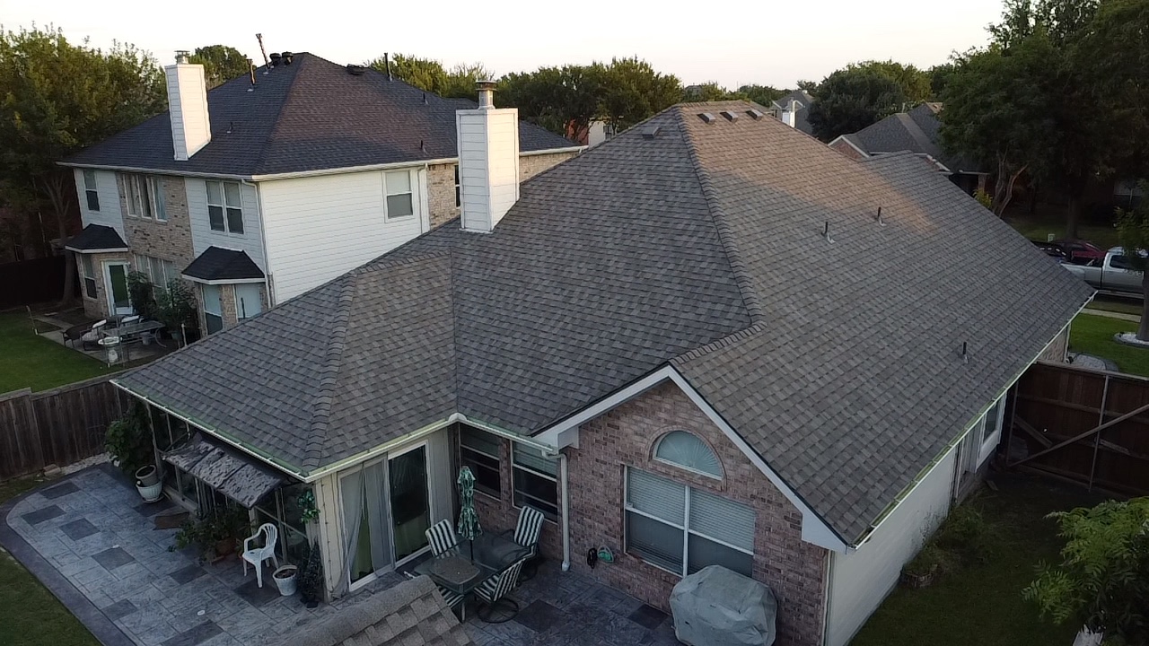 An aerial view of the roofs of suburban homes, with a focus on one house's backyard patio and sunroom. The patio features a table with chairs and an umbrella, a white plastic chair, and large potted plants. The sunroom is visible with its glass doors and windows. The surrounding yards are green and lined with trees and fences. Several vehicles are parked in the background.