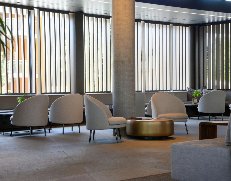 A stylish commercial lounge featuring light-colored rounded tub chairs arranged on a gray tiled floor, a central gold drum coffee table, a large concrete structural pillar, and vertical window louvers.