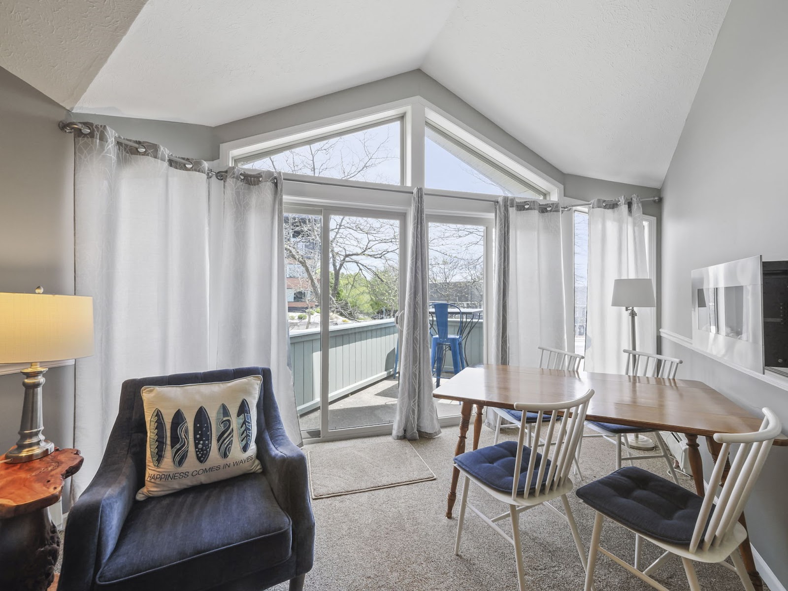 A bright and airy dining and living area with a vaulted ceiling, large windows, and a balcony. The room features a navy blue armchair with a "Happiness Comes in Waves" pillow, a wooden dining table with white chairs and navy cushions, and a floor lamp. The balcony has a small table and chairs and a view of trees and buildings.