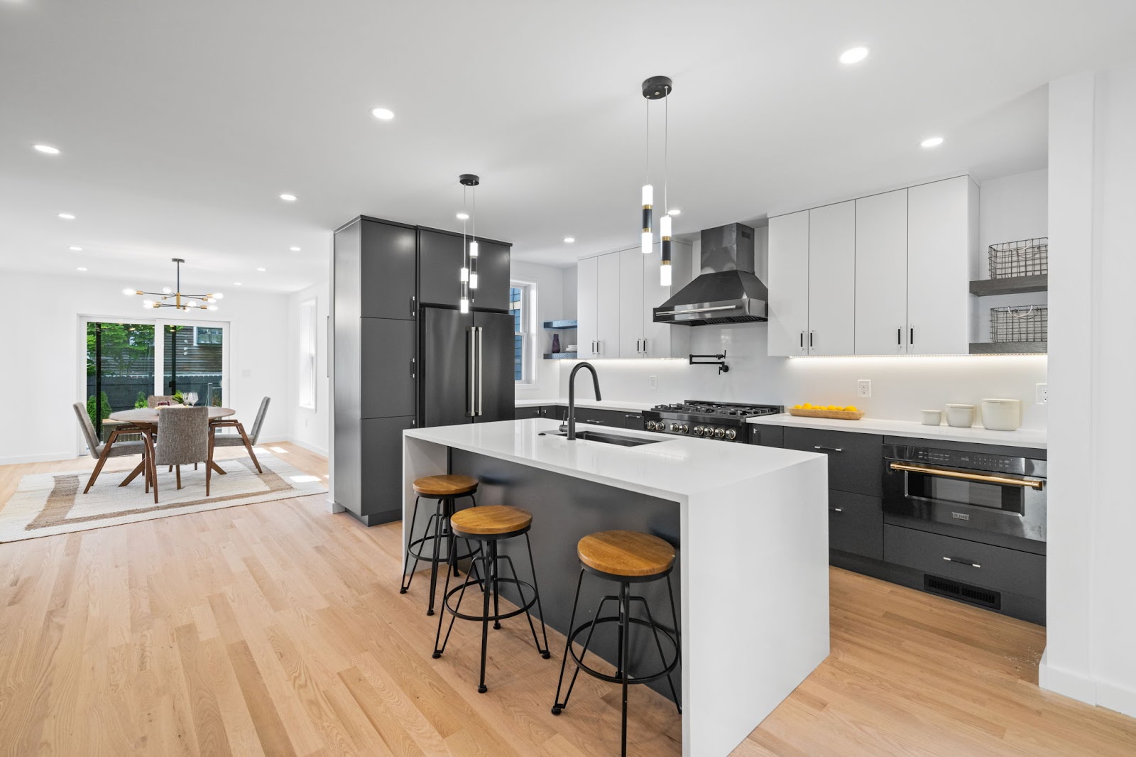 A modern kitchen and dining area with light wood floors, dark gray and white cabinetry, a large island with pendant lighting, and a dining table near a sliding glass door.