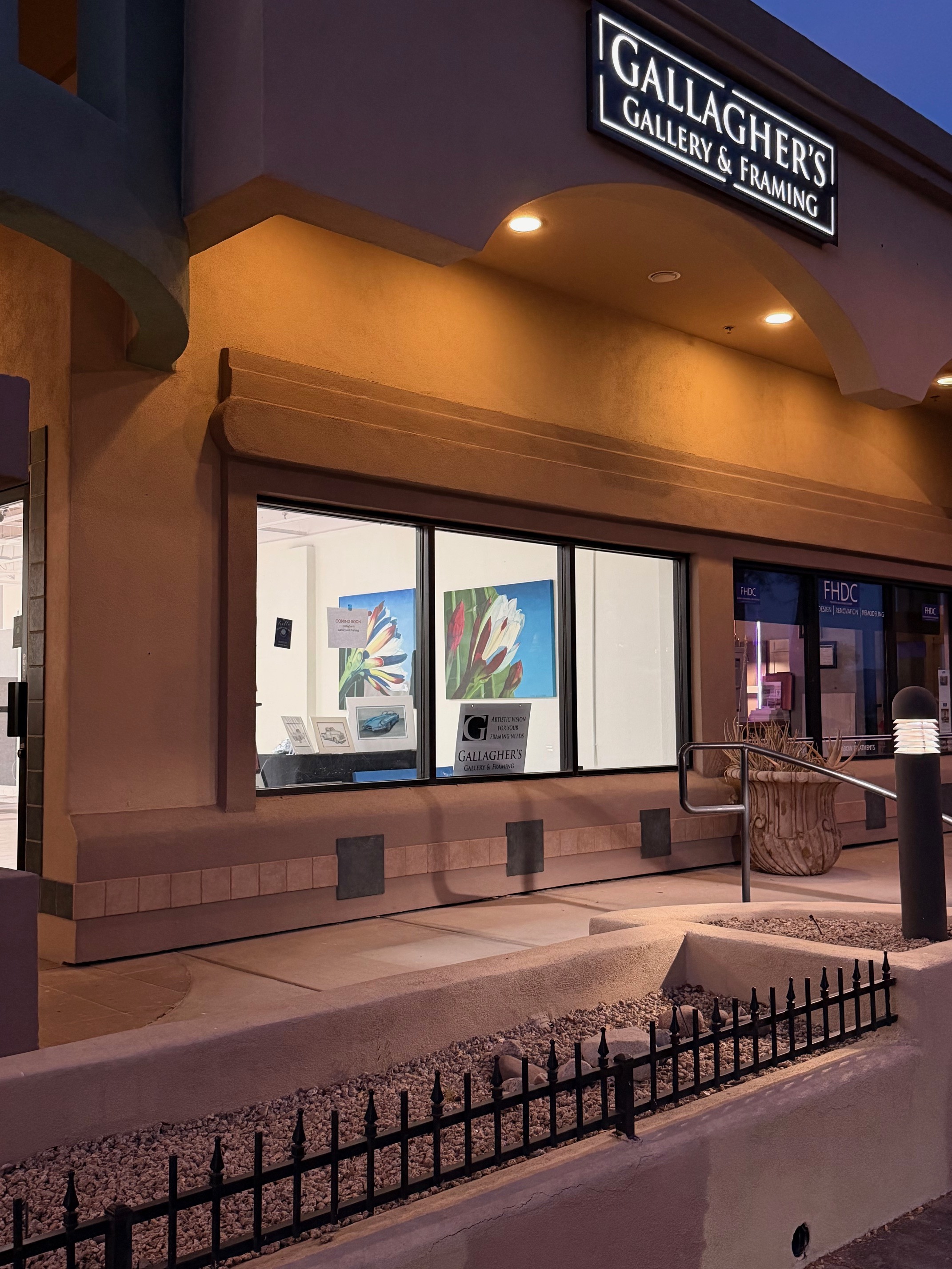 Exterior view of Gallagher&rsquo;s Gallery & Framing storefront at dusk, featuring illuminated signage, large display windows with artwork, warm architectural lighting, and a clean, modern entrance with decorative elements.