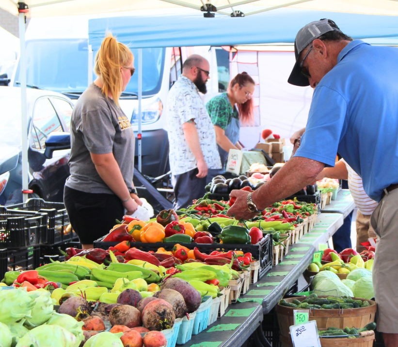 Shoppers browsing a colorful outdoor produce stand loaded with fresh bell peppers, deep purple beets, cabbages, and cucumbers under a blue canopy tent
