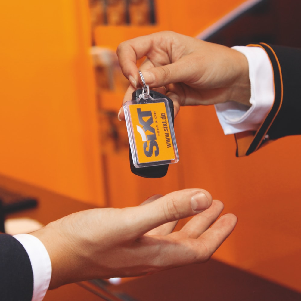 Ein Sixt-Mitarbeiter legt einem Kund*innen einen Schlüsselbund mit Sixt-Logo-Schlüsselanhänger in die wartenden Hände.