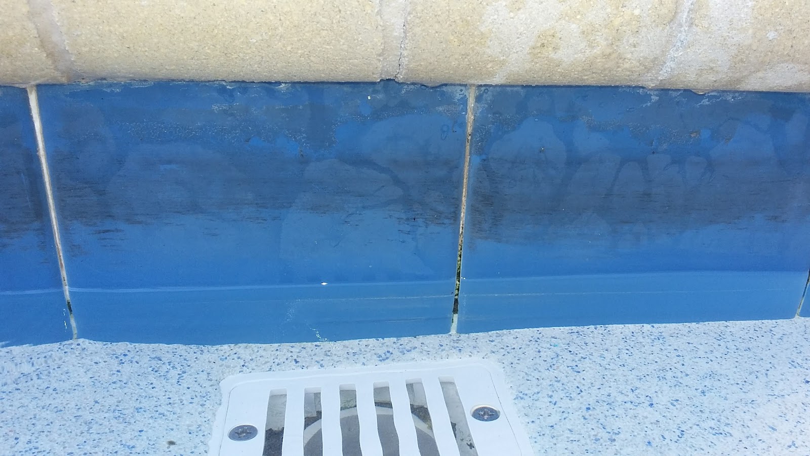Close-up of a swimming pool drain area with blue tile, waterline staining, and surrounding pool surface.