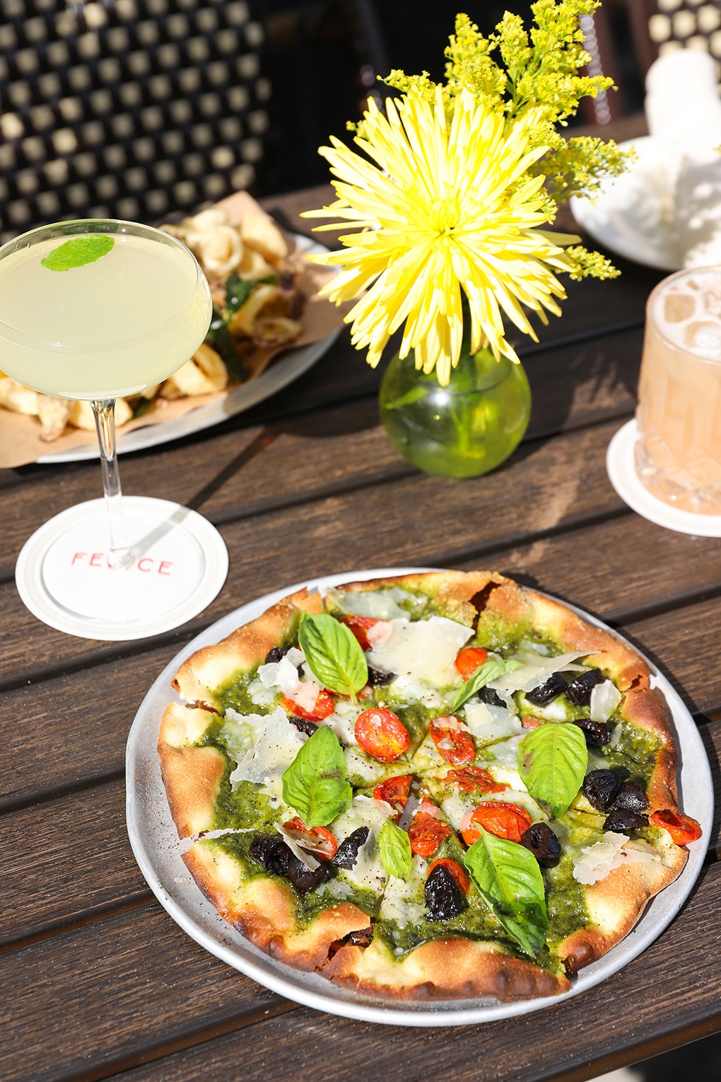 A bright, sunlit shot of a thin-crust pizza topped with pesto, cherry tomatoes, and olives next to a pale green cocktail and a yellow flower in a vase on a patio table.