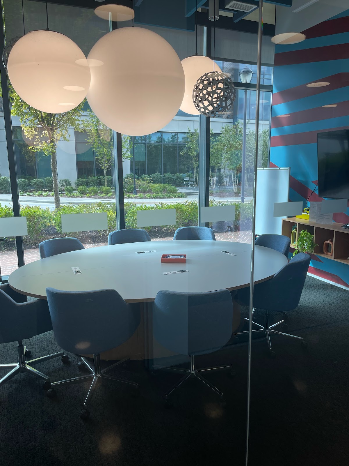A modern conference room with a large oval table, blue upholstered chairs, and large spherical pendant lights. The room has large windows overlooking a landscaped outdoor area with trees and buildings. The walls feature abstract geometric patterns in blue and red.