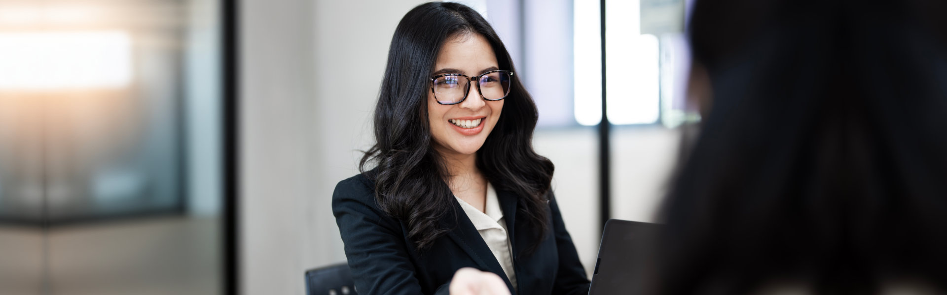 A smiling woman with dark, wavy hair and glasses is wearing a black blazer over a cream-colored blou 