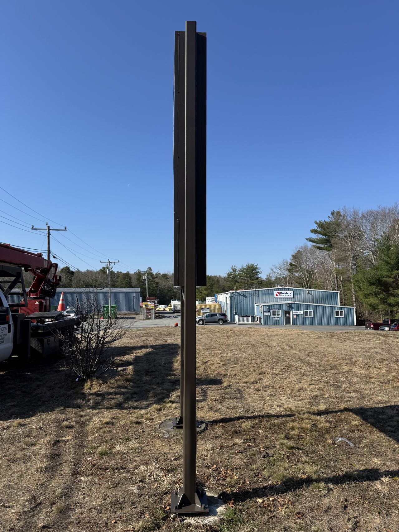 Side view of a tall Builders FirstSource sign structure with the store building visible in the background, showing the exterior grounds of a lumber and building materials facility.