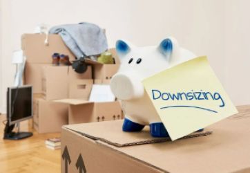 A classic white ceramic piggy bank with blue accents is positioned on a corrugated cardboard moving box. A yellow sticky note with the handwritten word "Downsizing" in blue is affixed to the piggy bank. In the blurred background, several other packed cardboard boxes, some with items spilling out, and a computer monitor are visible on the floor of a room.