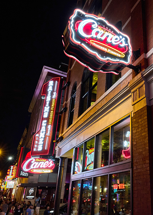 A low-angle night shot looking up at a brick building exterior adorned with bright red neon Raising Cane's signs, including a vertical vintage-style blade sign reading "Raising Cane's Est. 1996".