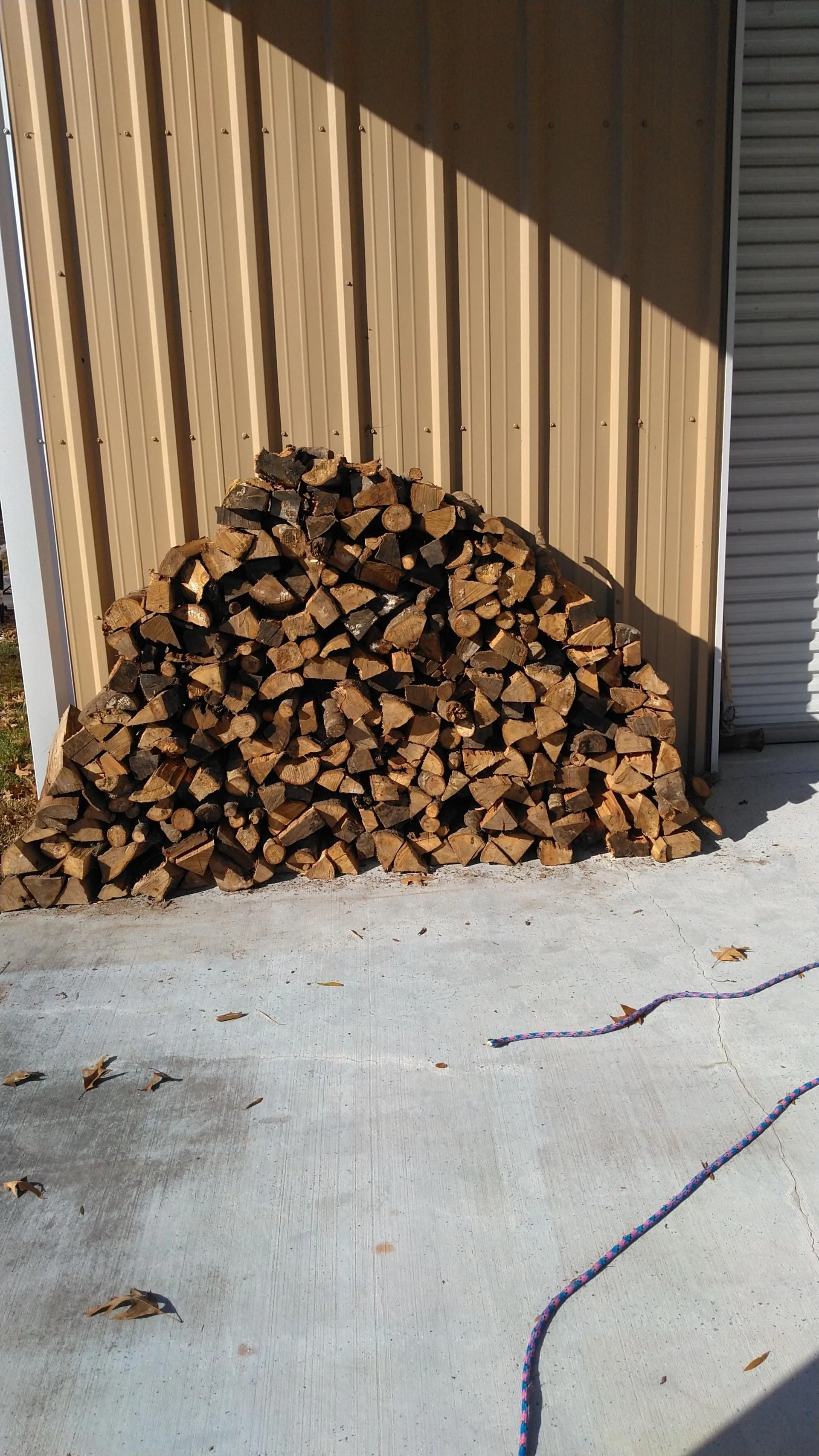 Neatly stacked firewood placed beside a tan metal building on a concrete surface, prepared for use during cold weather months.