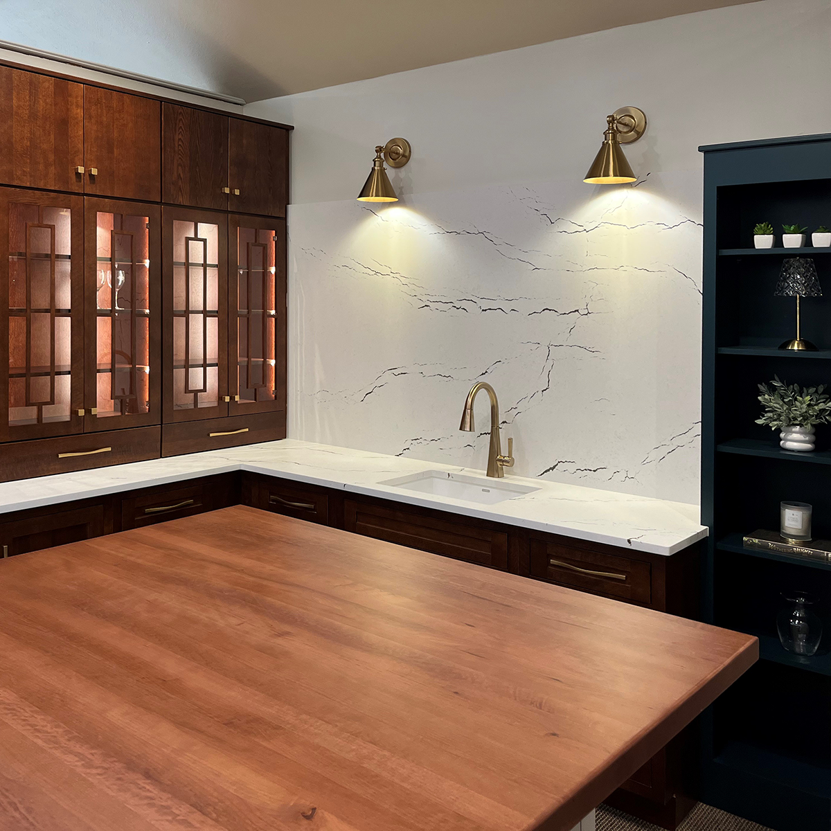 Close-up of a showroom kitchen featuring Decora Marquis dark wood cabinets, a large wood island countertop, white quartz backsplash with subtle veining, brass wall sconces, and a modern sink with a gold-tone faucet.