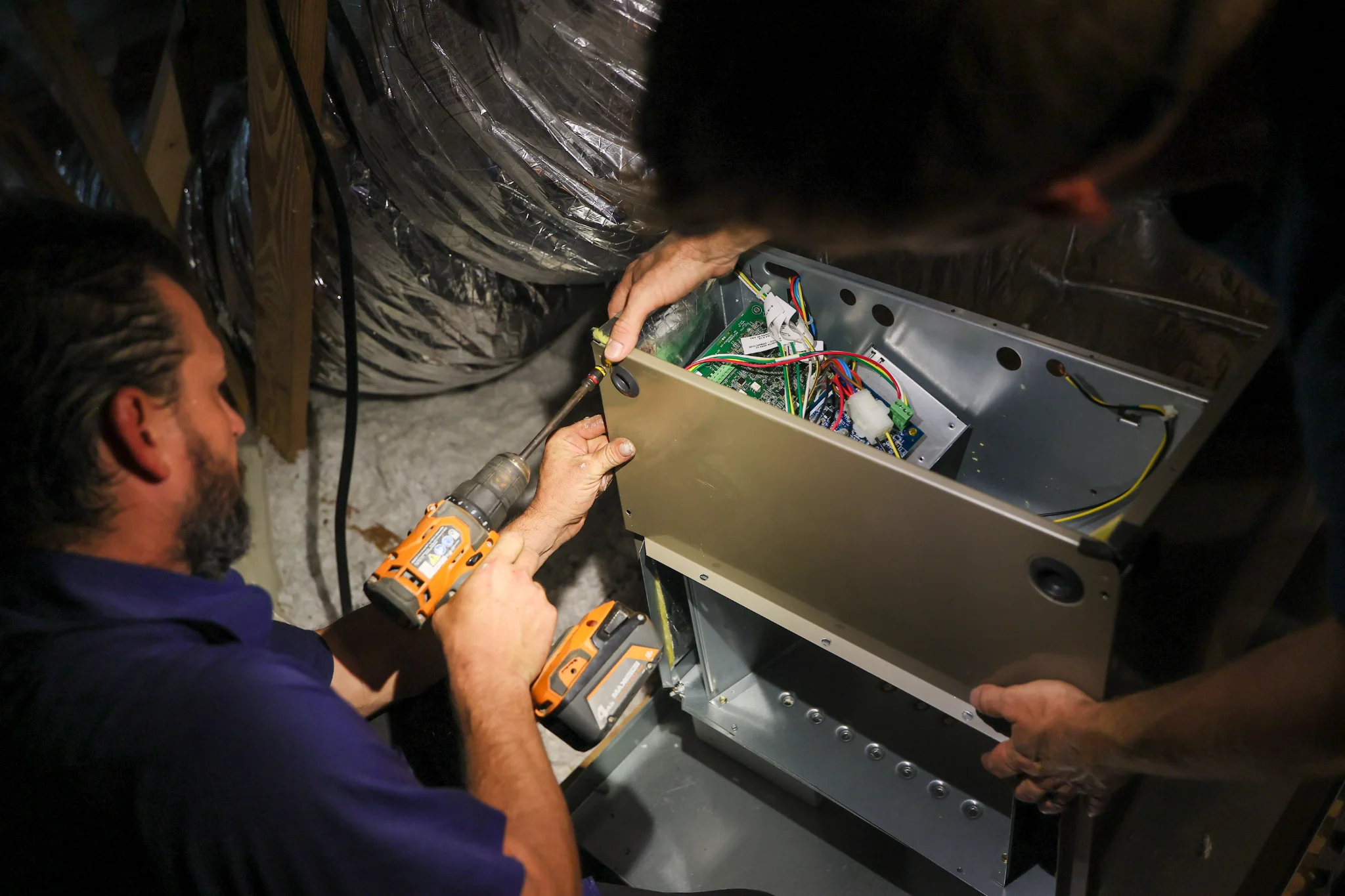Two men are working on an HVAC system in an attic. One man, wearing a purple shirt, is using a cordl 