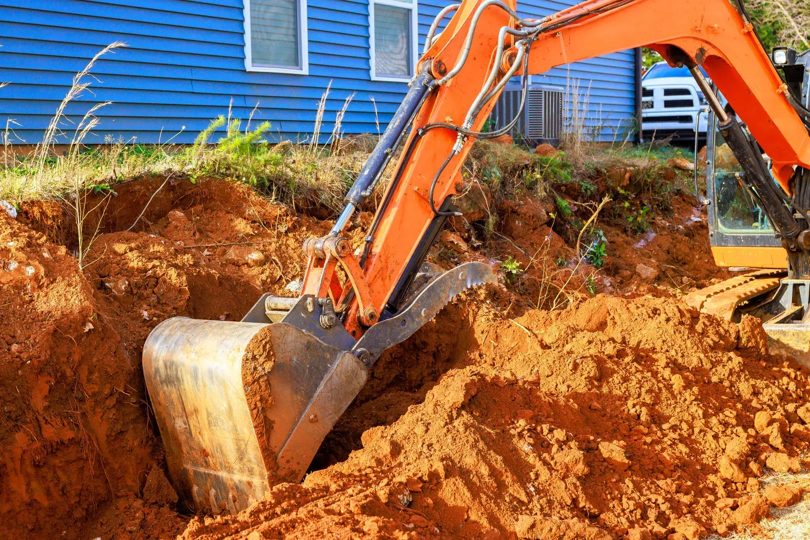 Excavator performing digging work near a residential property, removing soil for foundation or utility installation. Heavy-duty construction equipment ensures precise excavation for grading, trenching, and site preparation in residential and commercial projects.