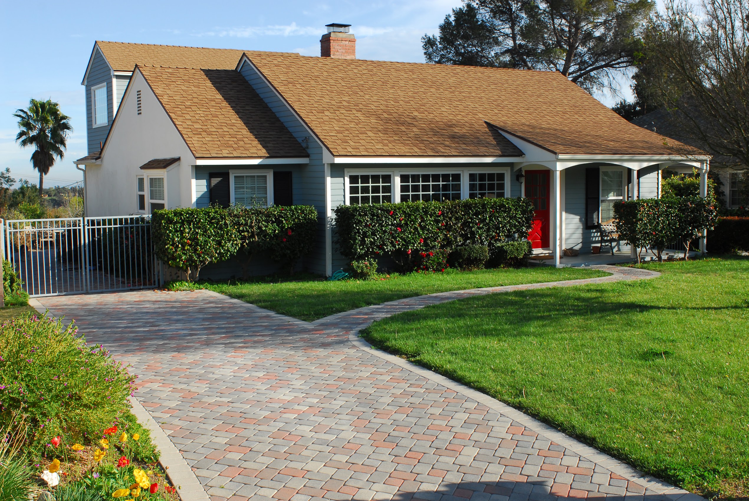 Sunny suburban cottage with a steep shingle roof, white siding, hedges, brick walkway, green lawn, palm trees, and blue sky.