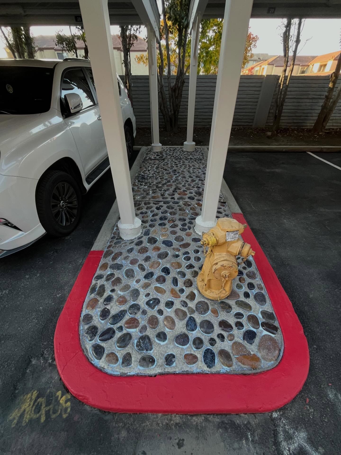 Decorative pebble-finished concrete pad surrounding a fire hydrant under a carport, bordered with red paint, showcasing functional hardscaping, drainage-friendly surface, and organized parking structure within a residential or apartment complex.