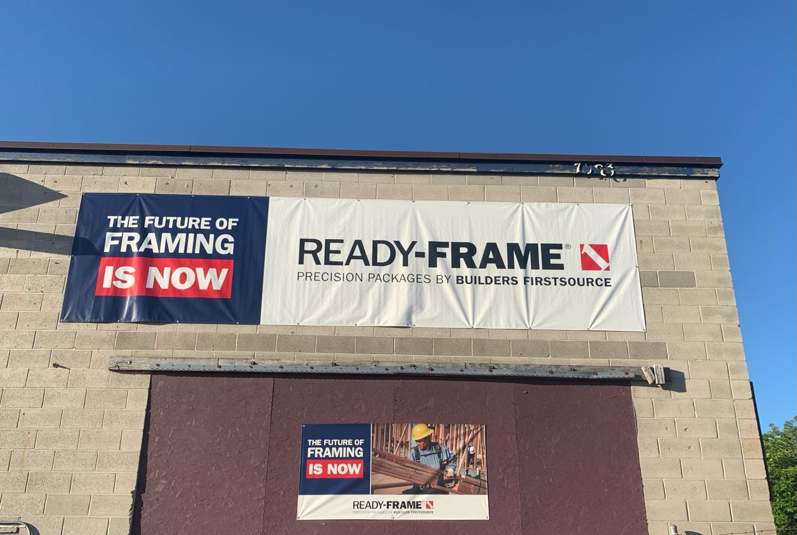 An exterior shot of the Builders FirstSource facility showcasing a large banner that reads 'The Future of Framing is Now' and 'READY-FRAME Precision Packages by Builders FirstSource.' The banner is mounted on a tan brick wall under a clear blue sky.