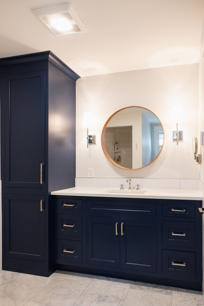 A modern bathroom vanity with dark navy cabinets, a white countertop, and a round wooden-framed mirror above the sink. Two wall-mounted sconces with white shades flank the mirror, and a tall, matching navy cabinet stands to the left. The floor is tiled with light gray marble.