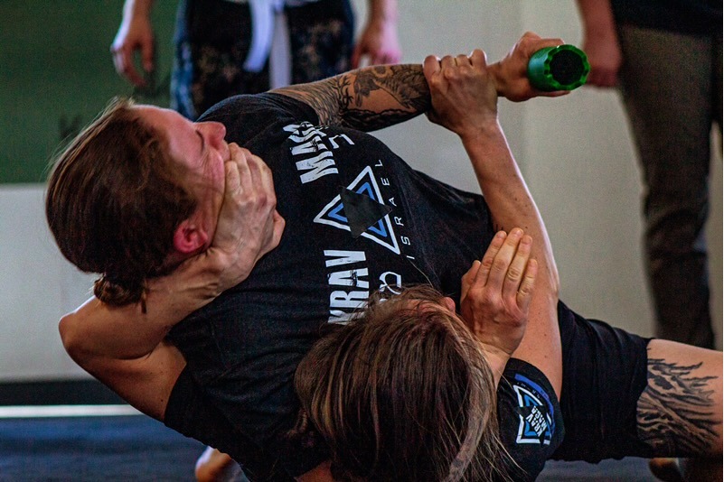 Two adult students working a close-range ground defense, one controlling the other's head and neck while they fight for control of a training weapon.