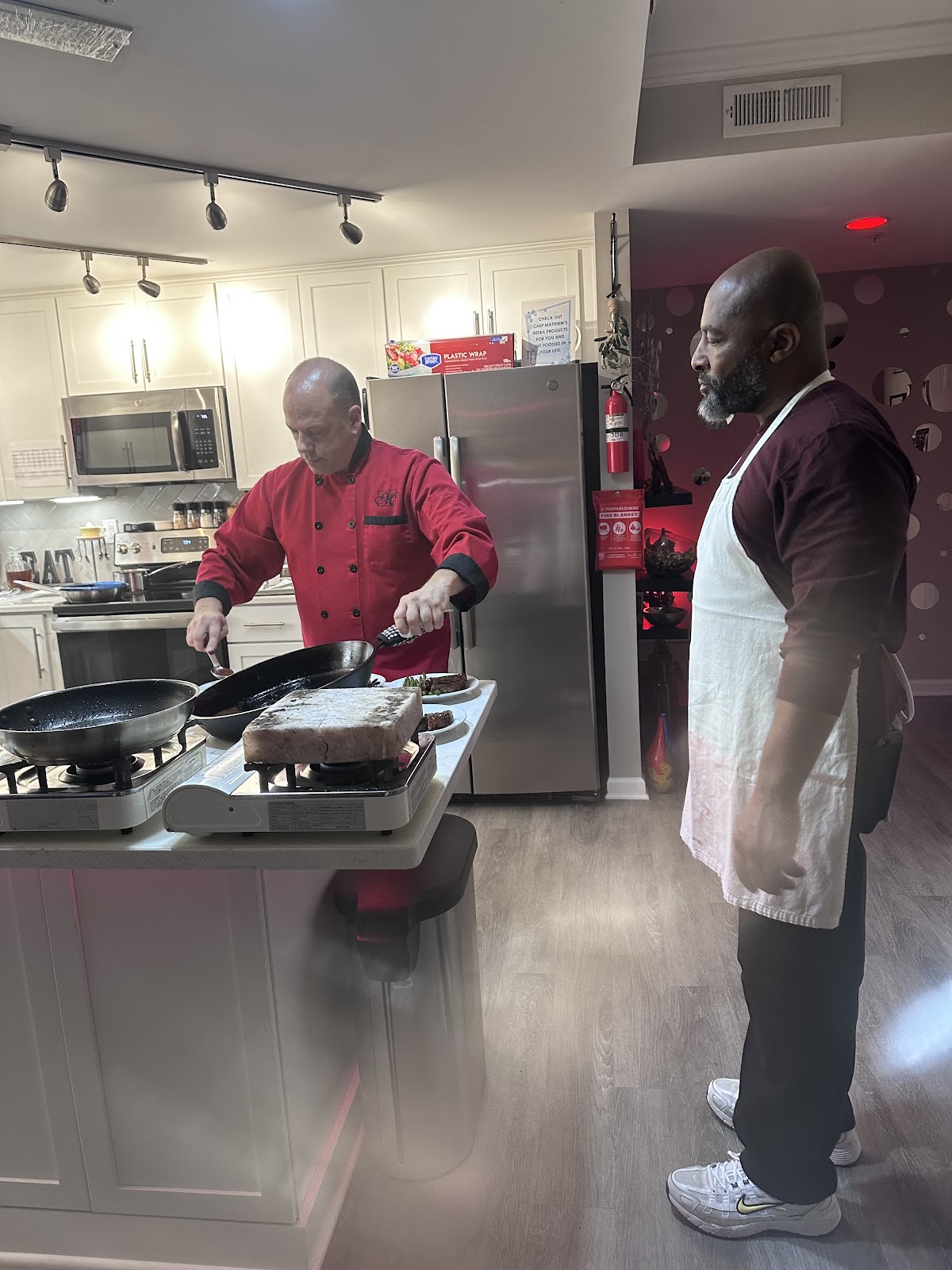 Two men are in a kitchen, one wearing a red chef's coat and the other an apron. The man in the red chef's coat is cooking on a portable stovetop with a pan and a block of food on it. There are plates with food, likely steaks, to the right of the stovetop. The second man stands to the right, observing the cooking. The kitchen appears to be modern with white cabinets and stainless steel appliances.