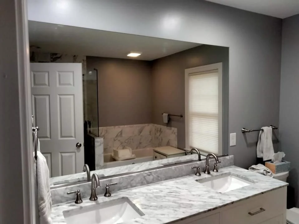 A modern bathroom featuring a double vanity with marble countertops, two sinks, and silver faucets, reflected in a large wall mirror. The mirror also reflects a white door, a glass-enclosed shower, and a bathtub with marble tiling. The walls are painted a neutral gray.