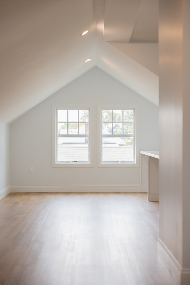 An empty room with a sloped ceiling, two white-framed windows looking out onto a residential area, and a light-colored wooden floor. A white table or counter is visible on the right side of the room. Recessed lighting fixtures are embedded in the ceiling.
