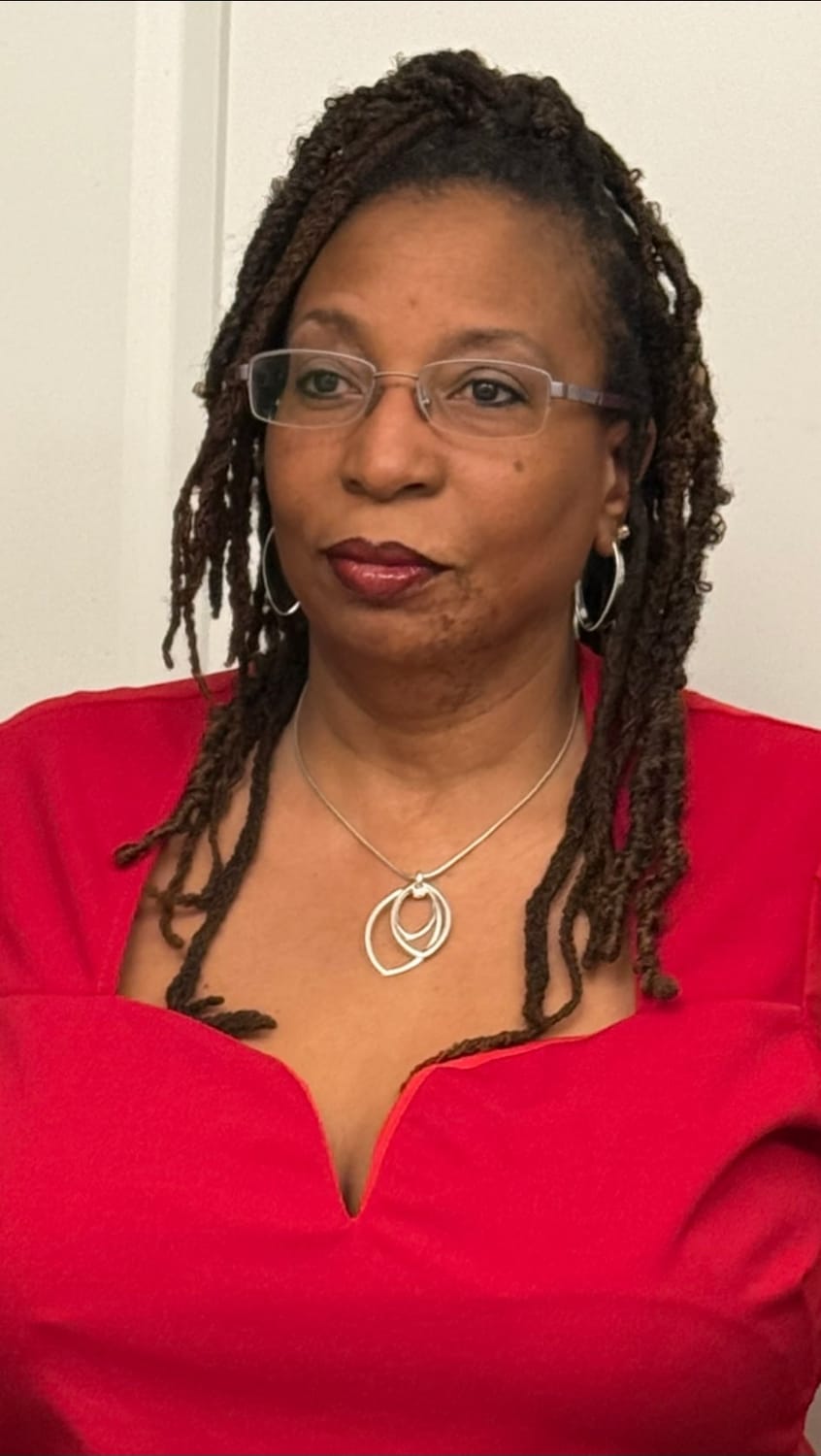 A portrait of a Black woman with dreadlocks and glasses, wearing a red V-neck dress and silver jewelry. She is looking to her left with a neutral expression. The background is a plain white wall.