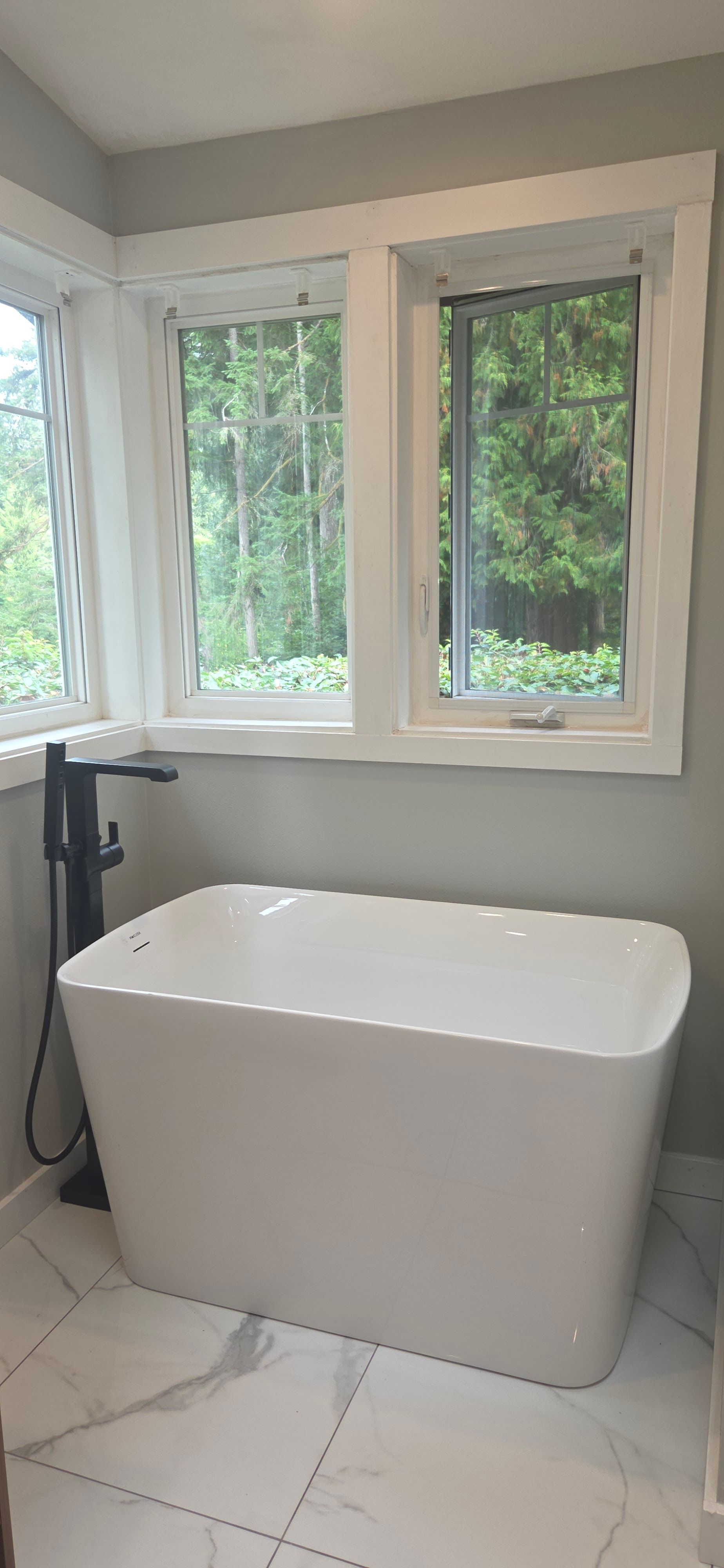 Modern freestanding bathtub installed beneath large windows with forest view, paired with a black fl 