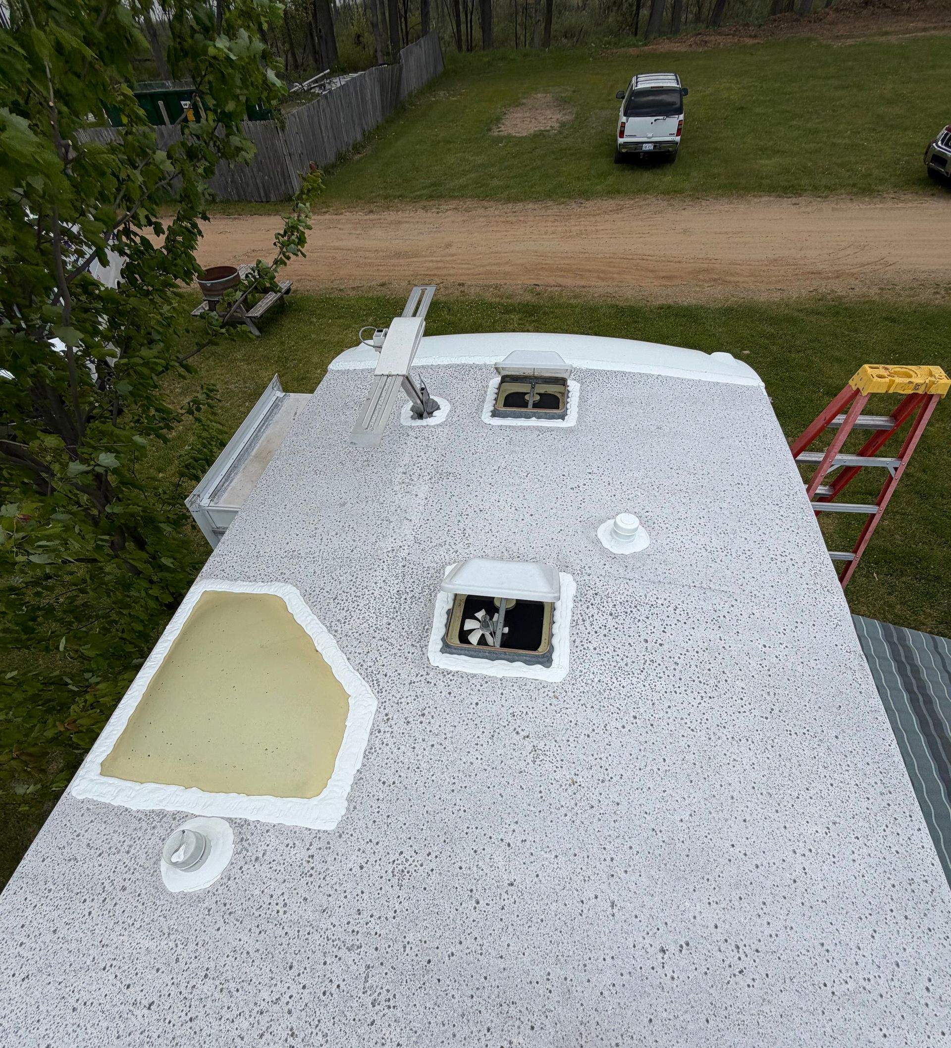 Top view of an RV roof with textured white coating, roof vents, sealed openings, antenna hardware, vent caps, and repair sealant, with a red ladder and parked vehicle visible nearby.
