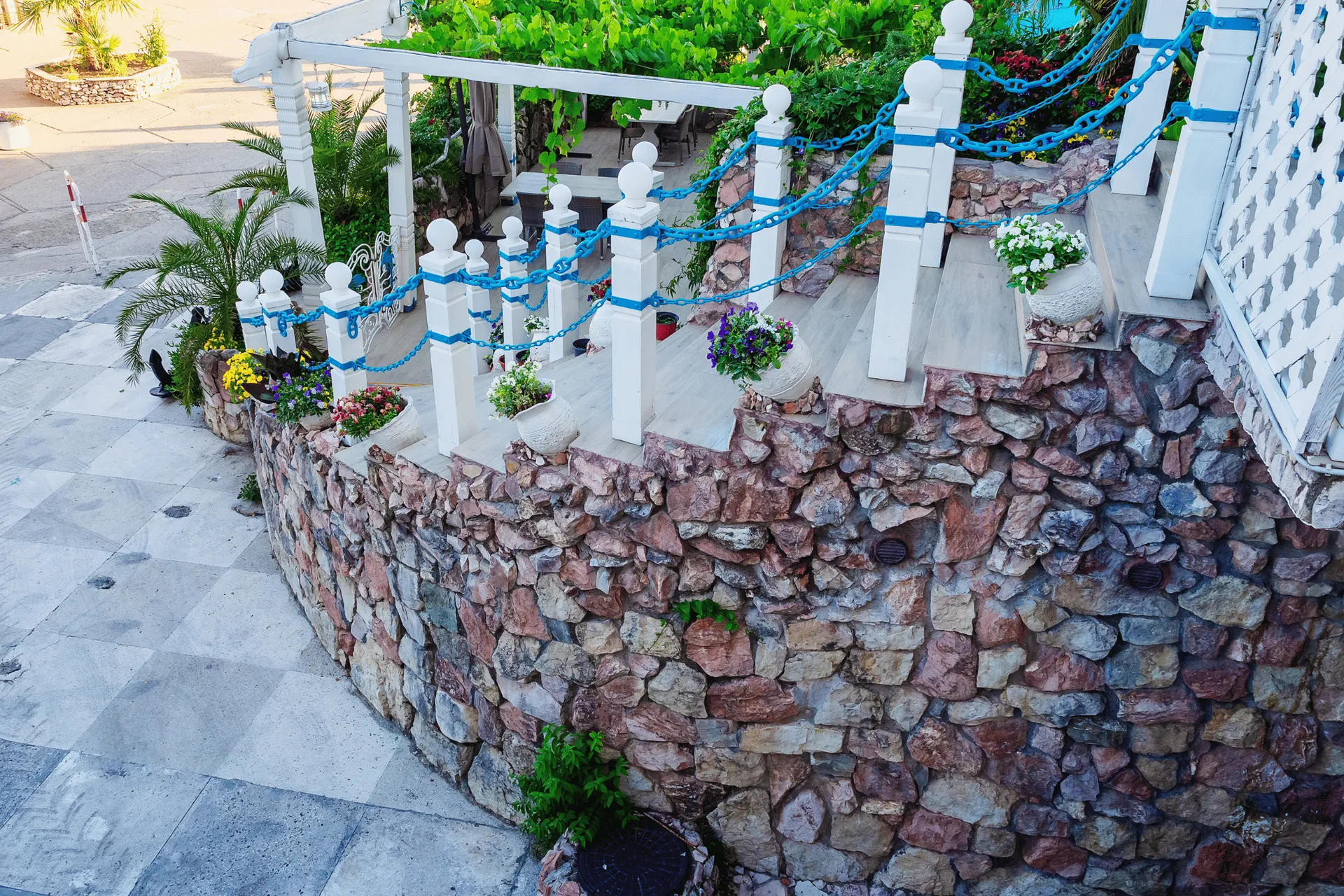 A curved stone wall with white posts and blue chain railings leads up a staircase adorned with potted plants. Lush green foliage drapes over the top of the structure.