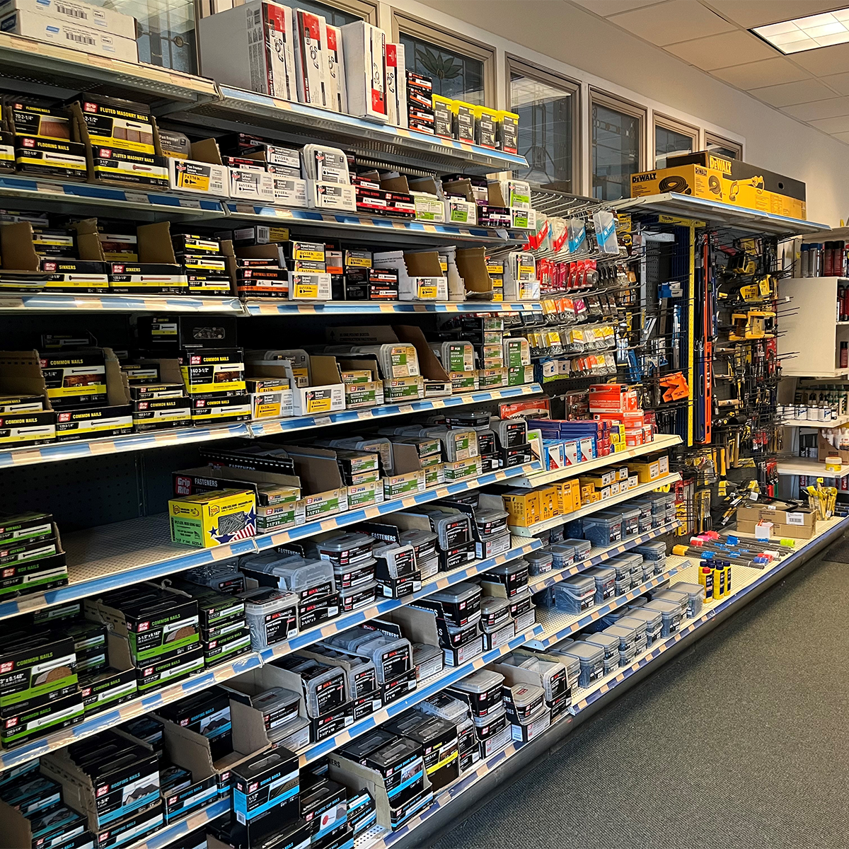 Retail hardware aisle stocked with fasteners, nails, screws, and anchors in labeled boxes and bins, with additional tools and accessories displayed on pegboards and shelves.