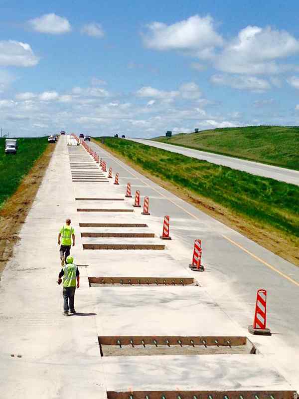 Eye-level view down a closed highway lane under clear blue skies, showing a long series of regularly spaced, rectangular slots cut into the concrete pavement for Dowel Bar Retrofit (DBR) installation. Two workers in high-visibility safety vests walk along the work zone past orange traffic control barrels.