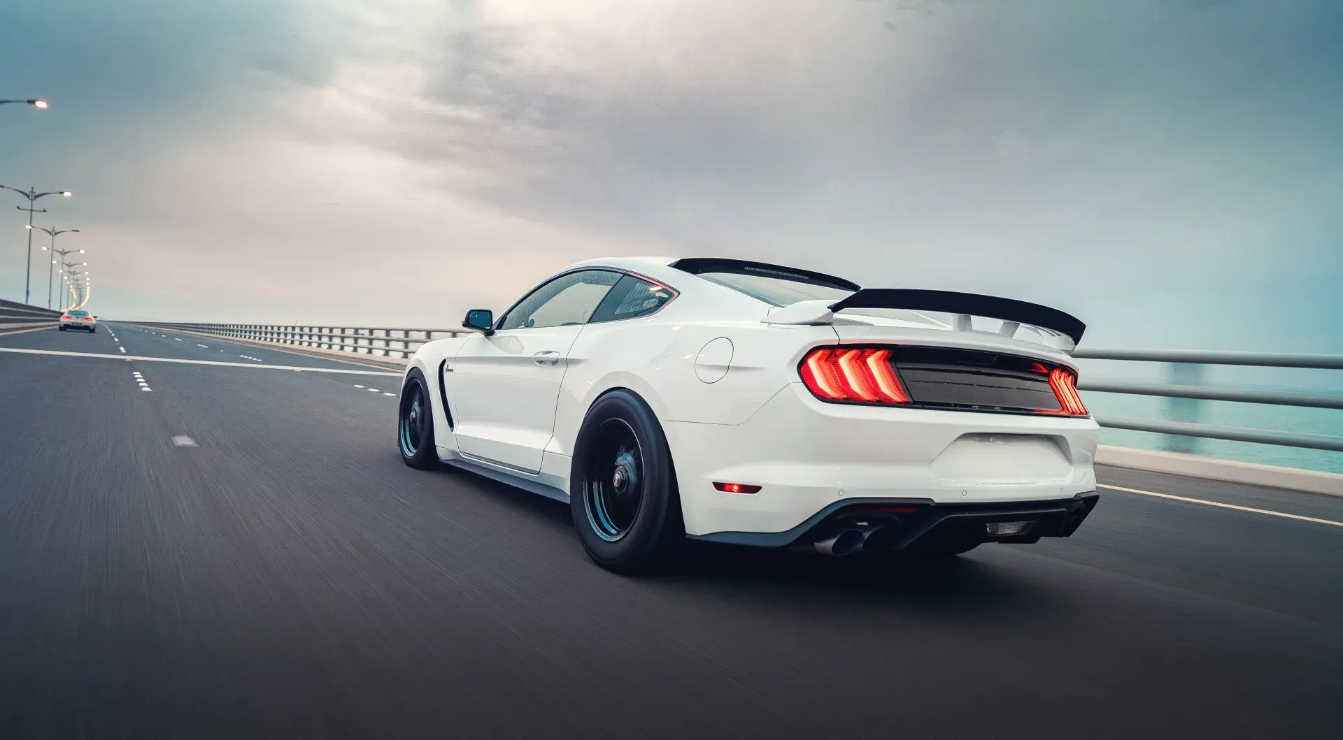 A white Ford Mustang GT500 is driving on a bridge with a blurred background of the ocean and cloudy sky. The car is captured from the rear-left, showcasing its sleek design and racing spoiler. The taillights are illuminated, and dual exhaust pipes are visible at the rear.