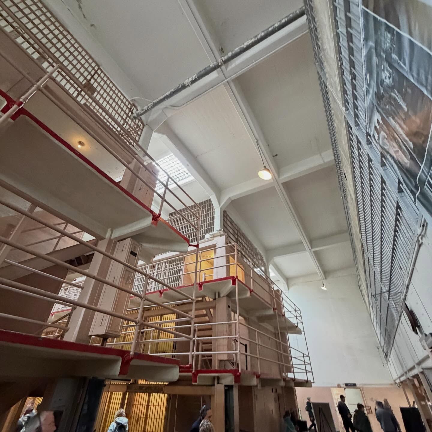 Interior view of a multi-level prison cell block featuring stacked tiers, metal railings, barred cells, and visitors exploring the facility, highlighting structural design, surveillance layout, and historical correctional architecture from a low-angle perspective.