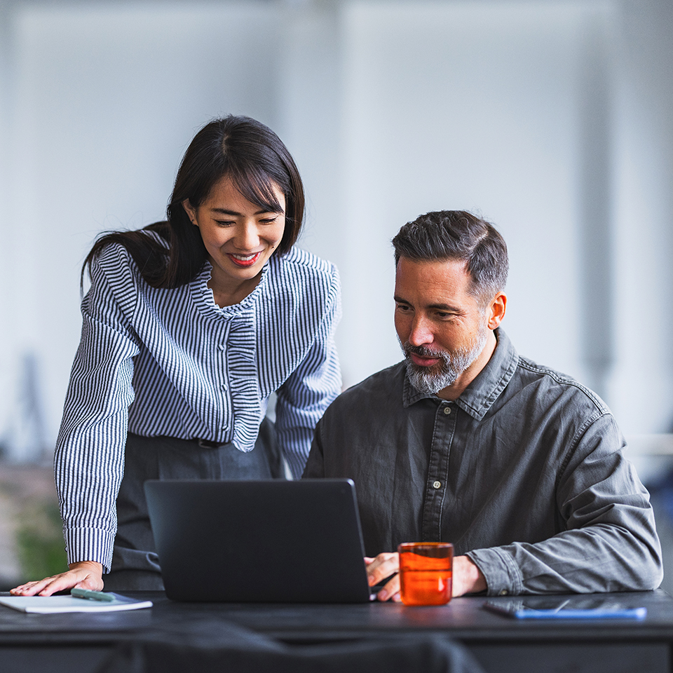 Two colleagues collaborate over a laptop at a desk in a modern office setting.