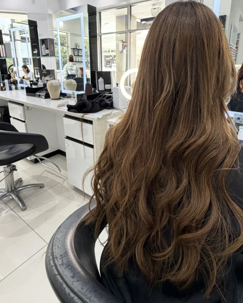 A person's long, wavy brown hair is prominently displayed from behind in what appears to be a hair salon. The background shows a clean, modern salon interior with mirrors, styling stations, hair products, and other salon equipment. Two women are visible in the background, one at a styling station and another reflected in a mirror.