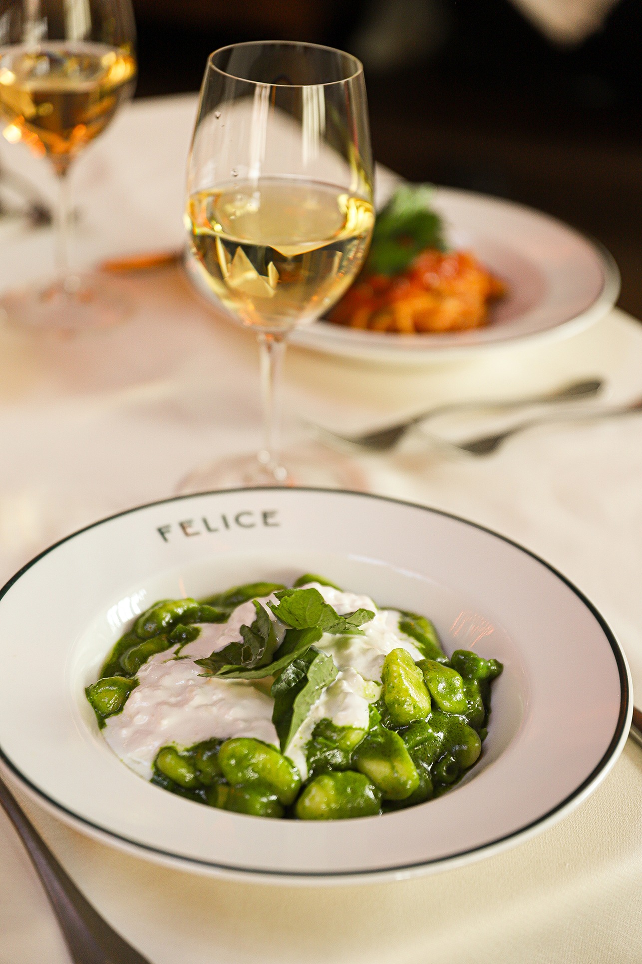 Soft gnocchi coated in a vibrant green pesto sauce and topped with creamy white burrata and fresh basil. A glass of crisp white wine sits in the soft-focus background.