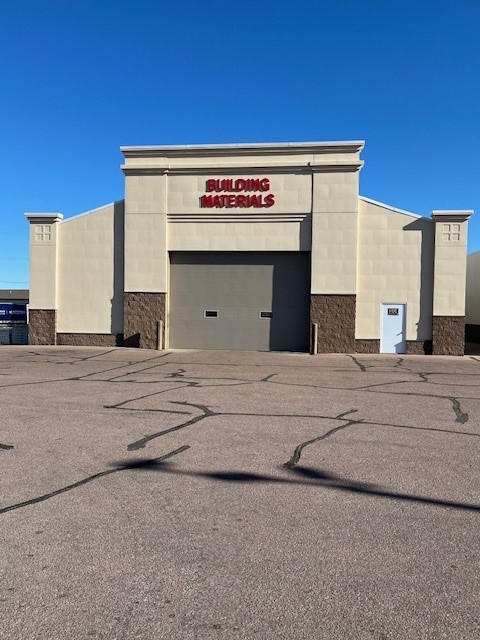 A beige building with a large sign reading "Building Materials" in red lettering is displayed. The structure features a central roll-up door and a smaller side door, with a mix of beige and brown tones on the facade. The parking lot in the foreground is empty, with visible cracks in the pavement. The scene is under a clear blue sky.