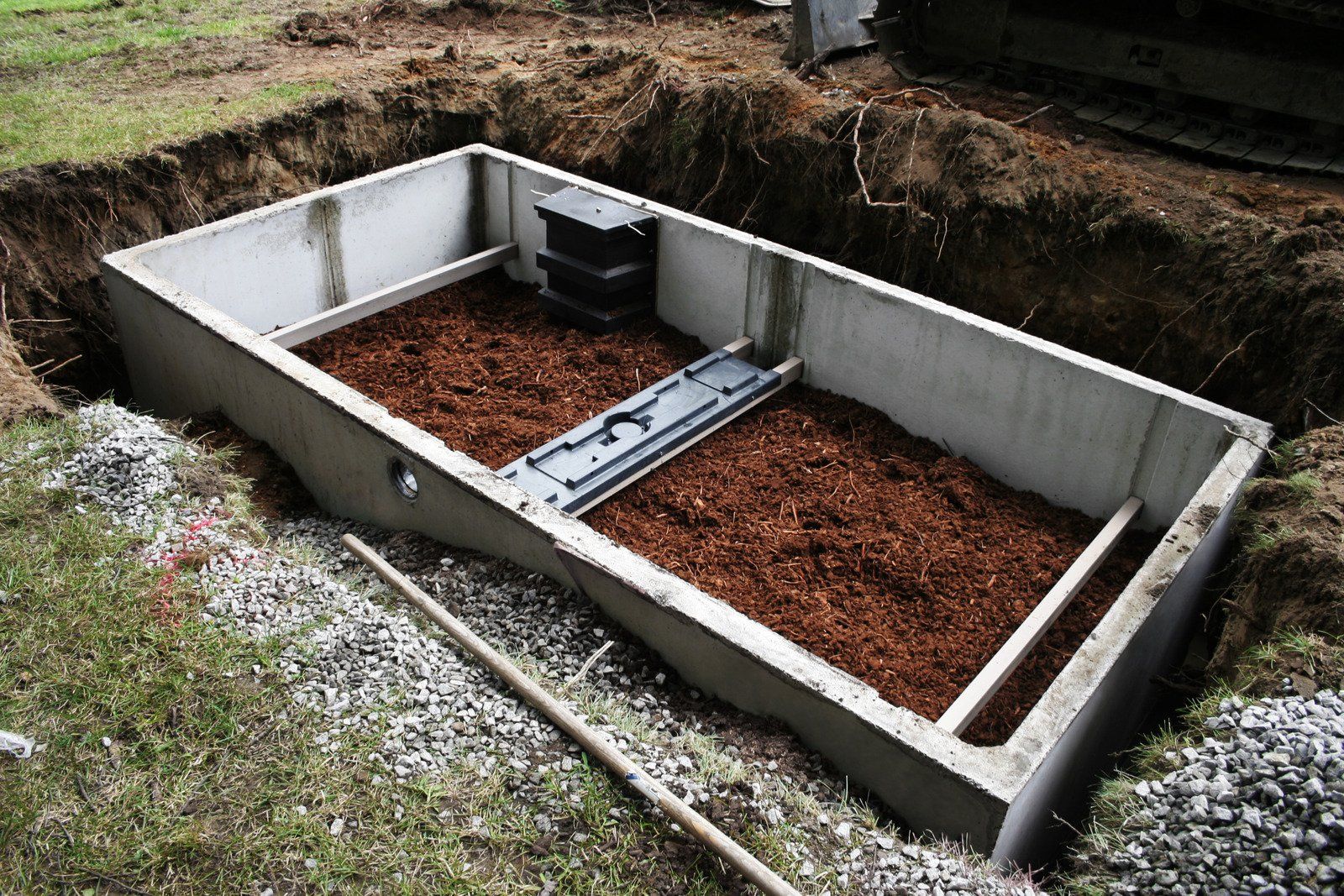 A large rectangular concrete septic tank sits partially buried in an excavated dirt pit. Inside, dark brown filtration material is visible alongside black plastic internal components and stabilizers.