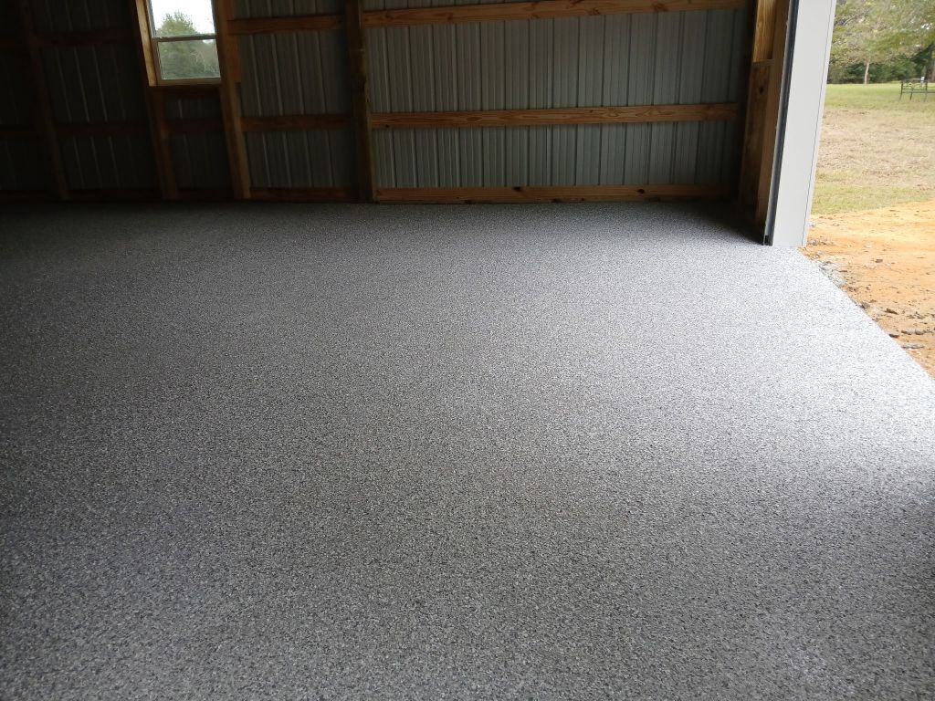 Interior of metal barn or garage featuring newly finished speckled epoxy concrete floor, wooden framing, corrugated wall panels, small window, and open doorway leading to grassy outdoor area.