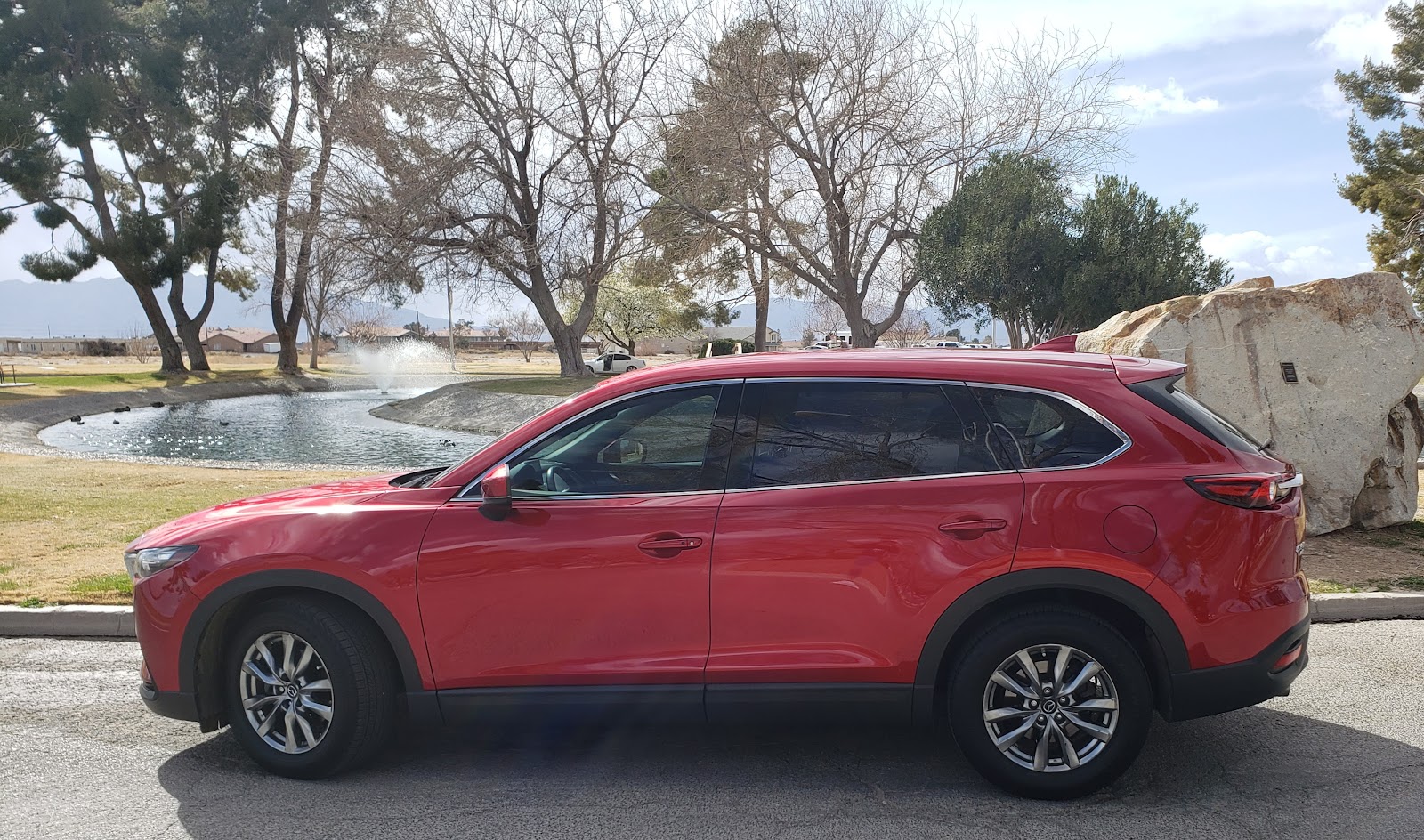 A red Mazda CX-9 SUV is parked next to a large boulder in a park-like setting. In the background, th 