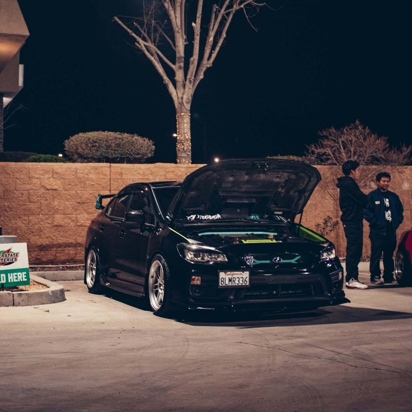 Modified black Subaru WRX displayed at a nighttime automotive gathering with its hood open, showcasi 