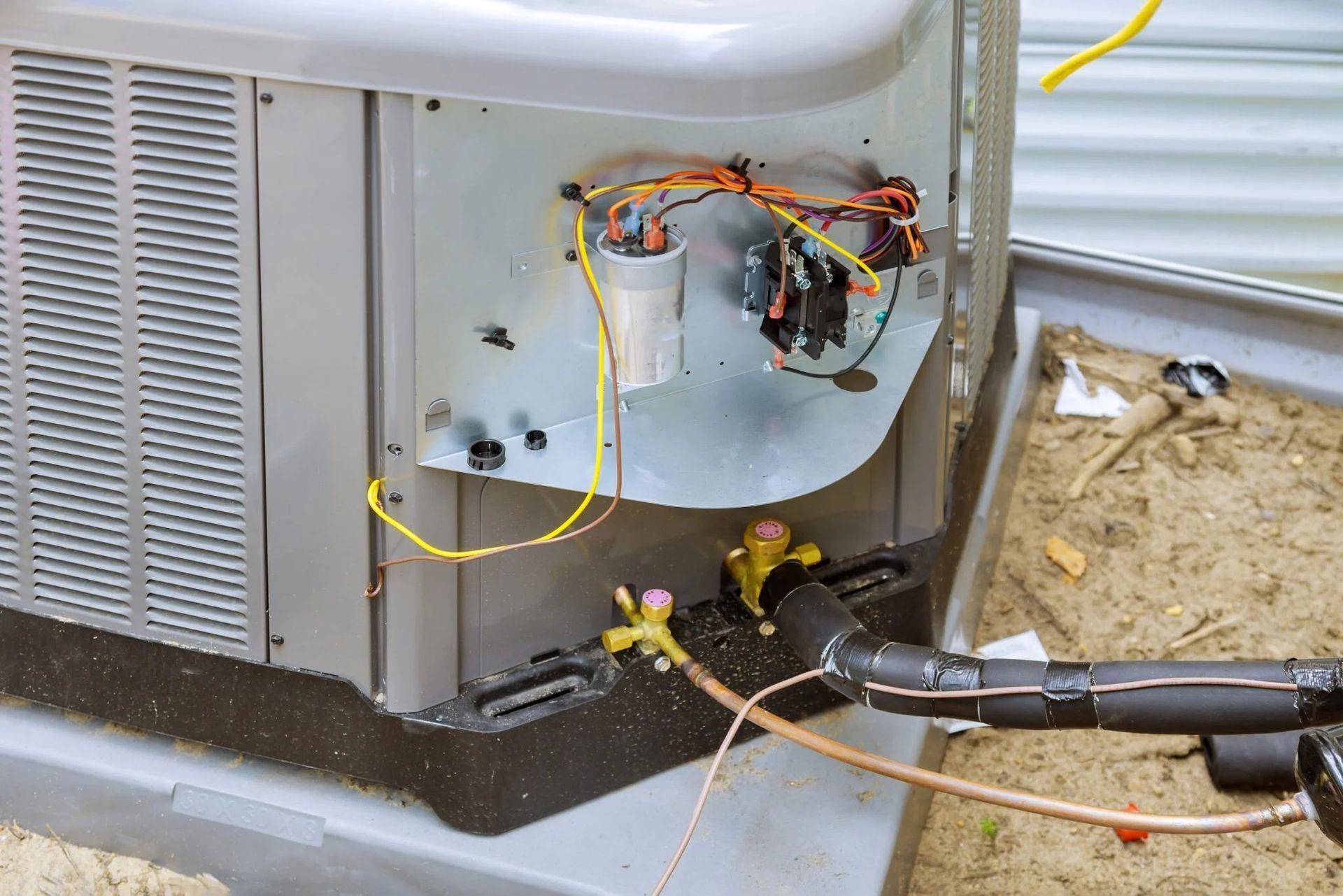 Outdoor air conditioning unit with exposed electrical components, capacitor, wiring, and refrigerant lines during maintenance or repair, showing internal HVAC system connections and service setup on residential condenser system.