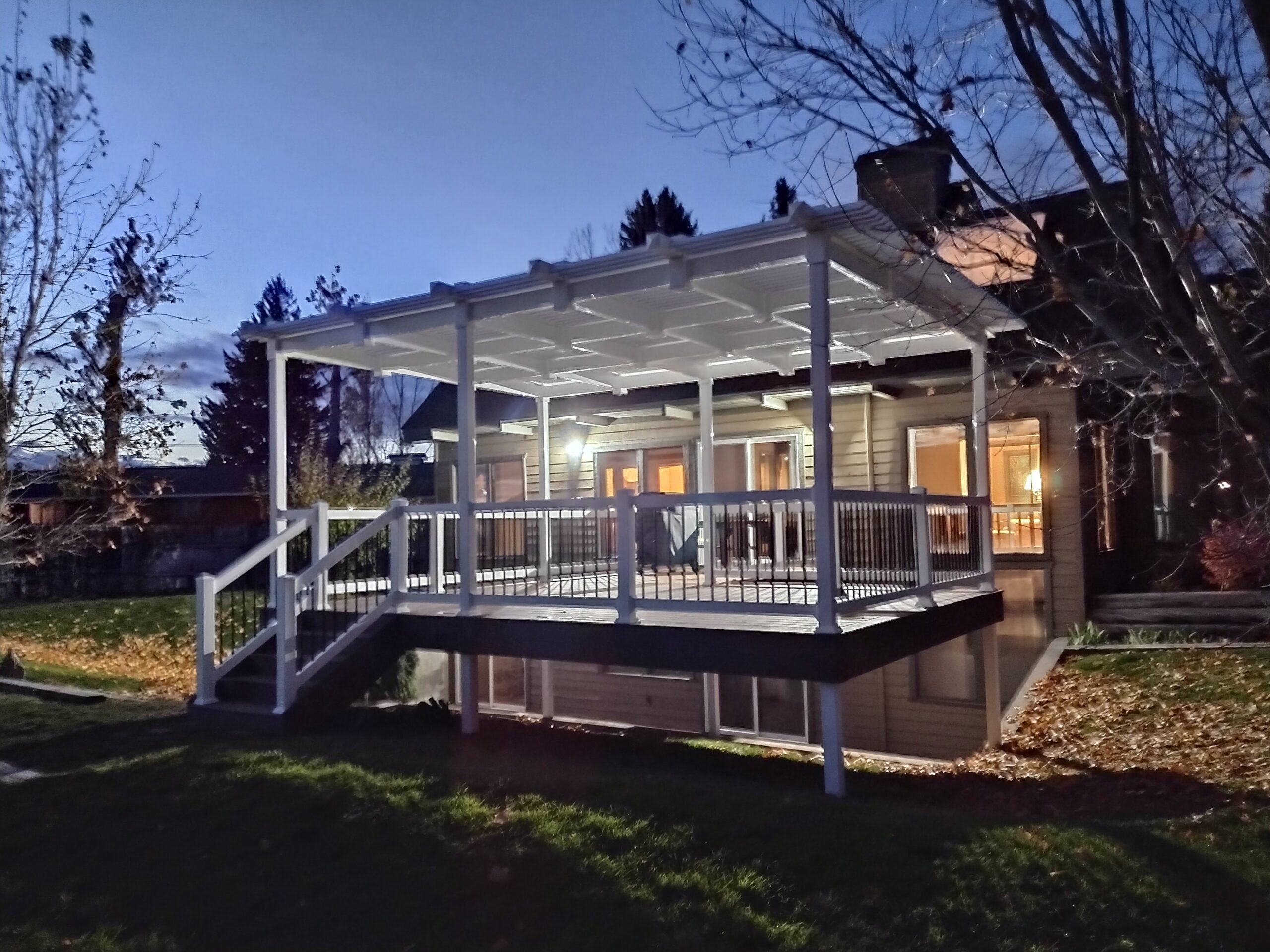 Extend your outdoor enjoyment well into the evening with a custom covered deck by DeckPro LLC. This raised composite deck features a solid patio cover for ultimate weather protection, allowing you to relax outside day or night. The stylish white railing with black balusters adds a modern touch to this cozy backyard retreat, illuminated by the house lights.