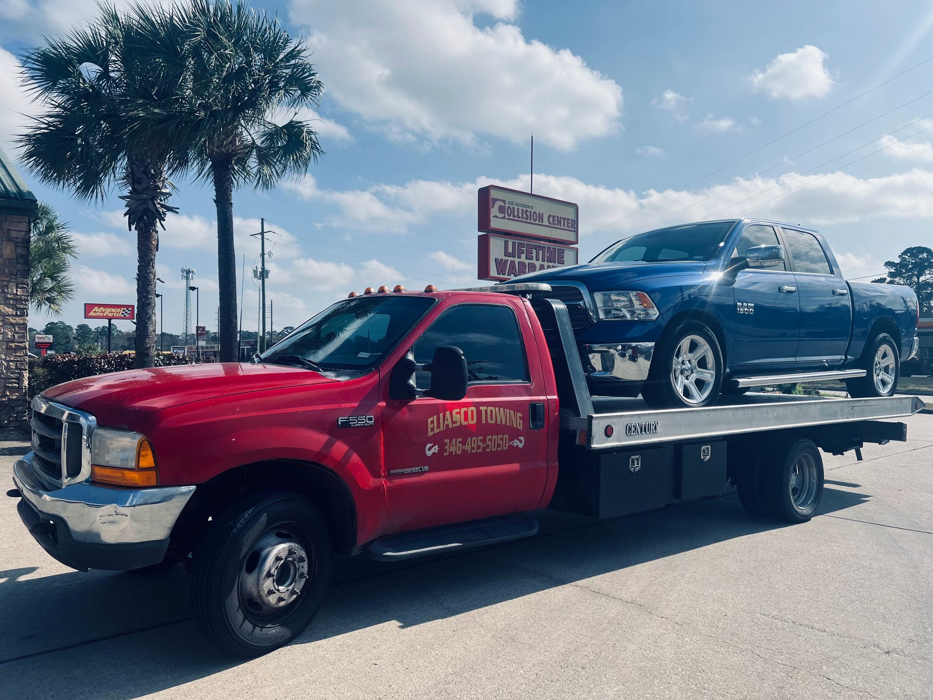 Eliasco Towing red flatbed truck transports a dark blue pickup outside an auto collision center, demonstrating professional vehicle recovery, towing, and transportation services in daylight.