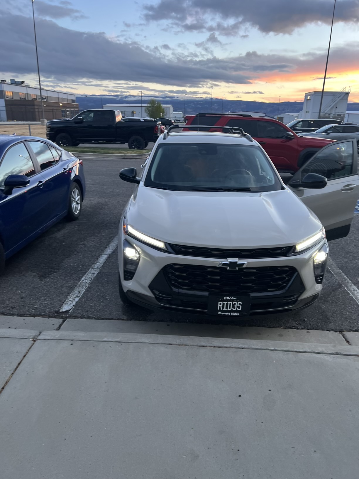 A white Chevrolet SUV is parked in a lot at sunset, with its driver's side door open. The SUV has a license plate that reads "RID3S" and "Elevate Rides". A blue sedan is parked to the left of the SUV, and a black pickup truck and a red SUV are parked in the background. The sky is a mix of gray clouds and vibrant orange and yellow hues from the sunset, with mountains visible on the horizon.