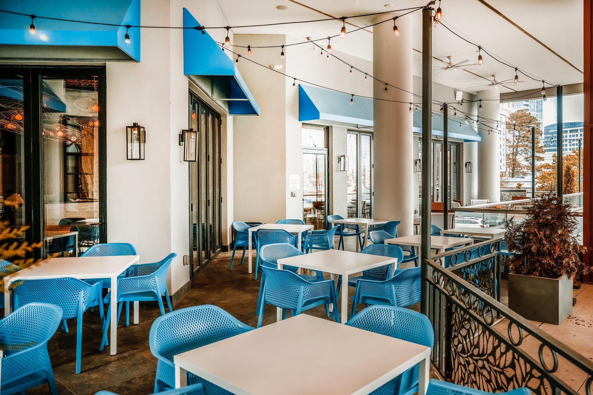 The patio area is lively and inviting, with a modern aesthetic characterized by vibrant blue chairs and sleek white tables. String lights add a warm, festive ambiance.